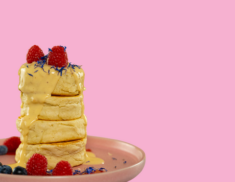 Turm aus fluffigen, saftigen Pancakes, mit Himbeeren, Blaubeeren und essbaren Blumen dekoriert. Dazu eine helle Toppingsauce, vor einem rosa Hintergrund.