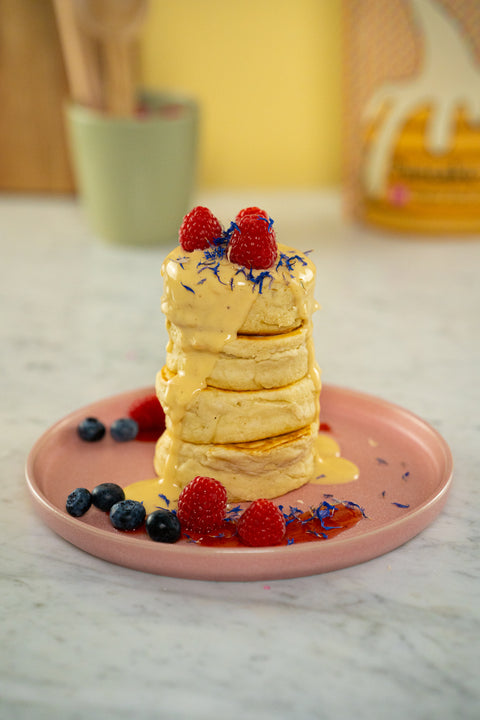 Turm aus super dicken, fluffigen Pancakes, garniert mit Himbeeren, Blaubeeren, essbaren Blumen und einer hellen Topping-Sauce. Im Hintergrund ist die Pancake-Mix-Tüte von Reoat zu erahnen.