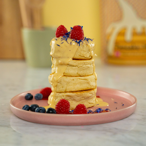 Turm aus fluffigen, saftigen Pancakes, mit Himbeeren, Blaubeeren und essbaren Blumen dekoriert. Dazu eine helle Toppingsauce, vor einem rosa Hintergrund.