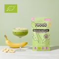reoat Banana Matcha Haferdrinkpulver als Bio-Getränkepulver in der grünen Packung „reoat BANANA MATCHA HAFERDRINKPULVER“ neben einem cremigen Banana-Matcha-Latte im Coupe-Glas (grün, schaumig, mit Eis), getoppt mit Bananenscheiben; daneben frische Banane und Bananenscheiben auf hellem Marmortisch vor grünem Hintergrund – veganes reoat Latte-Pulver zum schnellen Anrühren (nur Wasser) für Banana Matcha Latte, Iced Matcha, fruchtige Kaffee-Alternative und reoat Drinks-Kollektion.