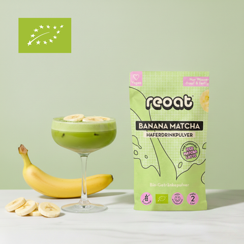 reoat Banana Matcha Haferdrinkpulver als Bio-Getränkepulver in der grünen Packung „reoat BANANA MATCHA HAFERDRINKPULVER“ neben einem cremigen Banana-Matcha-Latte im Coupe-Glas (grün, schaumig, mit Eis), getoppt mit Bananenscheiben; daneben frische Banane und Bananenscheiben auf hellem Marmortisch vor grünem Hintergrund – veganes reoat Latte-Pulver zum schnellen Anrühren (nur Wasser) für Banana Matcha Latte, Iced Matcha, fruchtige Kaffee-Alternative und reoat Drinks-Kollektion.
