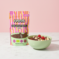 reoat Breakfast Bowl Kakao als Bio-Hafermischung („reoat BREAKFAST BOWL KAKAO“, „Nur Wasser drauf & fertig“) neben einer Schale cremigem Schoko-Porridge/Breakfast Bowl, getoppt mit Bananenscheiben, Schokodrizzle/Schokosauce, Kakaonibs oder Schokostückchen und frischen Himbeeren; Szene auf hellem Marmortisch vor rosé Hintergrund – vegane reoat Frühstücksbowl zum schnellen Anrühren nur mit Wasser, ideal für Frühstück, Snack oder Meal-Prep, schokoladige Bowl für Bowl-Lover und Fans von Kakao-Porridge.