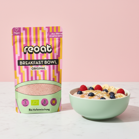 reoat Breakfast Bowl Original als Bio-Hafermischung („reoat BREAKFAST BOWL ORIGINAL“, „Nur Wasser drauf & fertig“) neben einer Schale cremiger Porridge/Breakfast Bowl, angerührt und getoppt mit frischen Beeren (Himbeeren, Blaubeeren), Bananenscheiben und Samen/Topping; Szene auf hellem Marmortisch vor rosé Hintergrund – veganes reoat Frühstück zum schnellen Anrühren nur mit Wasser, ideal für gesundes Frühstück, Snack oder Meal-Prep, glutenfrei-geeignet je nach Variante und perfekt für Bowl-Lover.
