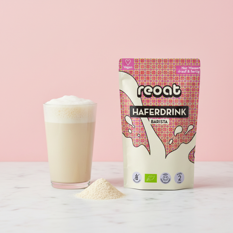 reoat Haferdrinkpulver Barista als Bio-Getränkepulver/Hafermilchpulver (200g für ca. 2 Liter) in der Packung „reoat HAFERDRINK BARISTA“ neben einem Glas fertig angerührtem Haferdrink mit cremigem Schaum; davor ein kleiner Haufen Haferdrinkpulver auf dem Marmortisch, Szene vor rosé Hintergrund – veganes reoat Oat Milk Powder zum schnellen Anrühren nur mit Wasser, ideal für Kaffee, Cappuccino, Latte, Müsli, Frühstück und als glutenfrei-geeignete Alternative (je nach Rezeptur).