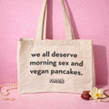 reoat Jutebeutel/Shopper aus naturfarbener Bio-Baumwolle mit großem Statement-Print „we all deserve morning sex and vegan pancakes.“ und reoat Logo; freigestelltes Produktfoto vor pinkem Hintergrund, unten dekoriert mit Perlenkette, Schmuck, Dattel und Blüte – nachhaltige reoat Tragetasche für Einkauf, Uni, Alltag und Festival, passend zur reoat Extras-Kollektion und als Geschenk für Fans von veganem Frühstück, Pancakes und Brunch.