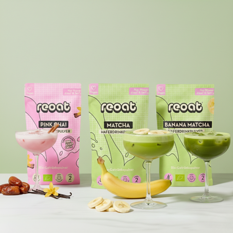 reoat Produktbild der Bio Latte-Getränkepulver als Set: Pink Chai Pulver (rosa Drink im Glas, mit Zimtstange), Matcha Haferdrinkpulver (grüner Matcha Latte im Coupe-Glas) und Banana Matcha Haferdrinkpulver (grüner Drink mit Bananenscheiben); vor den Packungen liegen Banane und Bananenscheiben, Datteln, Vanilleschote und Blüte – vegane, bio-zertifizierte, glutenfreie reoat Latte-Pulver zum schnellen Anrühren (nur Wasser) für Matcha Latte, Pink Chai Latte und Banana Matcha, ideal für Kaffee-Alternative, Iced 