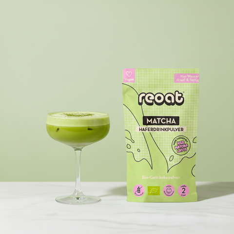 reoat Matcha Haferdrinkpulver als Bio-Getränkepulver in der grünen Packung „reoat MATCHA HAFERDRINKPULVER“ neben einem cremigen Matcha Latte/Matcha Drink im Coupe-Glas (grün, leicht schaumig, mit Eisstücken) auf hellem Marmortisch vor grünem Hintergrund – veganes reoat Matcha-Pulver/Latte-Pulver zum schnellen Anrühren (nur Wasser) für Matcha Latte, Iced Matcha, Kaffee-Alternative und Drinks-Kollektion.