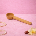 reoat Produktbild eines Holz-Messlöffels (Portionierer/Dosierlöffel) für Pulver und Mixe, liegend auf rosa Untergrund; im Hintergrund sind Haferflocken, ein goldener Löffel, Perlenkette sowie feines grünes Pulver zu sehen – praktisches reoat Zubehör zum genauen Abmessen von Haferdrinkpulver, Latte-Pulvern (z. B. Matcha), Pancake- und Waffel-Mix oder Breakfast-Bowls/Poridge, ideal für schnelle Zubereitung ohne Waage.
