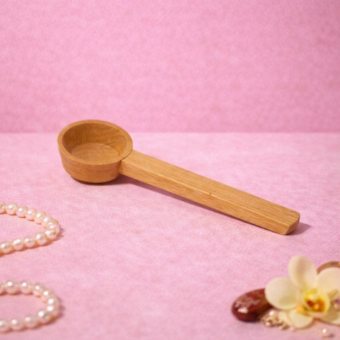 reoat Produktbild eines Holz-Messlöffels (Portionierer/Dosierlöffel) für Pulver und Mixe, liegend auf rosa Untergrund; im Hintergrund sind Haferflocken, ein goldener Löffel, Perlenkette sowie feines grünes Pulver zu sehen – praktisches reoat Zubehör zum genauen Abmessen von Haferdrinkpulver, Latte-Pulvern (z. B. Matcha), Pancake- und Waffel-Mix oder Breakfast-Bowls/Poridge, ideal für schnelle Zubereitung ohne Waage.
