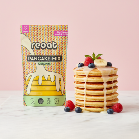 reoat Pancake-Mix Original als vegane Backmischung („reoat PANCAKE-MIX ORIGINAL“, „Nur Wasser drauf & fertig“) neben einem Stapel fluffiger veganer Pancakes mit cremigem Topping/Sirup, garniert mit Banane, Himbeeren, Blaubeeren und Minze auf Marmortisch vor rosé Hintergrund – schneller reoat Pfannkuchen-Teig zum Anrühren nur mit Wasser, ideal für Frühstück, Brunch und Familien-Backen, vegan ohne Milch und Ei aus dem reoat Onlineshop.
