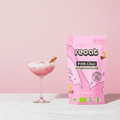 reoat Pink Chai Haferdrinkpulver als Bio-Getränkepulver in der pinken Packung „reoat PINK CHAI HAFERDRINKPULVER“ neben einem rosa Pink-Chai-Latte im Coupe-Glas mit Eis und cremigem Schaum, dekoriert mit Zimtstange; Szene auf hellem Marmortisch vor rosa Hintergrund – veganes reoat Latte-Pulver zum schnellen Anrühren (nur Wasser) für Pink Chai Latte, Iced Chai, süß-würzige Kaffee-Alternative und reoat Drinks-Kollektion.