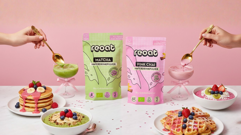 reoat Titelbild/Startseiten-Hero mit Bio Matcha Haferdrinkpulver und Bio Pink Chai Haferdrinkpulver in der Mitte; links ein grüner Matcha Latte im Glas und Pancakes mit pinkem Topping und Beeren, rechts ein rosa Pink Chai Latte im Glas sowie Waffeln und Breakfast Bowl mit frischem Obst (Himbeeren, Blaubeeren, Kiwi, Banane); zwei Hände toppen die Drinks mit goldenen Löffeln – pinkes Lifestyle-Set auf Marmortisch, vegane reoat Drinks und Frühstücksmischungen zum schnellen Anrühren (nur Wasser), ideal für Früh