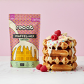 reoat Waffel-Mix Original als vegane Bio-Backmischung („reoat WAFFEL-MIX ORIGINAL“, „Nur Wasser drauf & fertig“) neben einem Stapel frisch gebackener Waffeln mit cremigem Topping/Glasur, bunten Streuseln, Bananenscheiben und Himbeeren; Streusel liegen auch auf dem Tisch, Szene auf Marmortisch vor rosé Hintergrund – veganer reoat Waffelteig zum schnellen Anrühren nur mit Wasser, ideal für Frühstück, Brunch oder Dessert, fluffige Waffeln ohne Milch und Ei aus dem reoat Onlineshop.