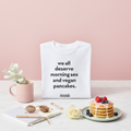 Produktbild eines weißen T-Shirts mit dem Spruch „we all deserve morning sex and vegan pancakes.“ und kleinem reoat Logo; Inszenierung auf Marmortisch mit rosa reoat Tasse, Notizbuch und Stift, Sonnenbrille, Schmuck, weißer Rose sowie einem Teller Pancakes mit Beeren und Sirup vor rosafarbenem Hintergrund – veganes Frühstücks-Lifestyle-Merch für reoat Extras/Kollektion.