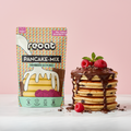 reoat Pancake-Mix Himbeer-Schoko als vegane Backmischung („reoat PANCAKE-MIX HIMBEER-SCHOKO“, „Nur Wasser drauf & fertig“) neben einem hohen Stapel fluffiger Pancakes mit Himbeer- und Schoko-Stückchen, überzogen mit reichlich Schokosauce/Schokoglasur und getoppt mit frischen Himbeeren sowie Minze; Schokoraspel und Stückchen als Deko auf dem Tisch, Szene auf Marmortisch vor rosé Hintergrund – veganer reoat Pfannkuchen-Mix zum schnellen Anrühren mit Wasser, ideal für Frühstück, Brunch oder Dessert, Raspberry-